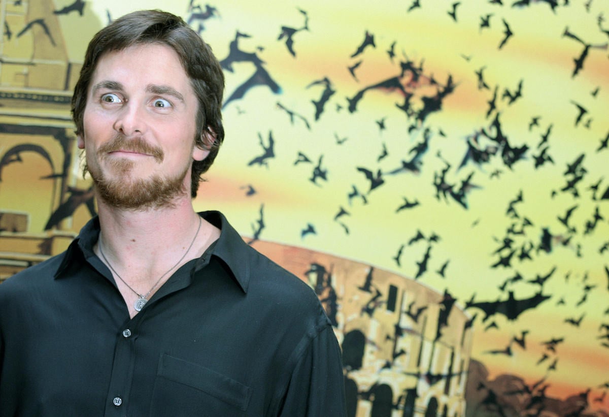 Christian Bale makes a wide-eyed expression as he stands in front of ‘Batman Begins’ artwork