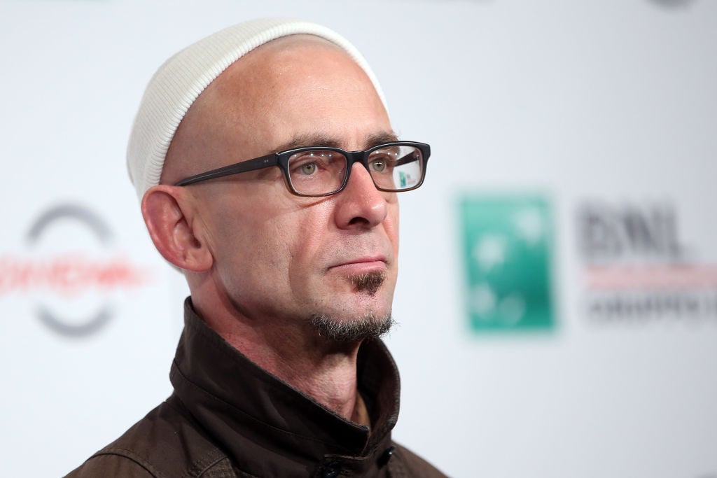 Chuck Palahniuk walks the red carpet in a dark suit and white cap. He wears glasses and looks out to the crowd of photographers with a sullen expression.