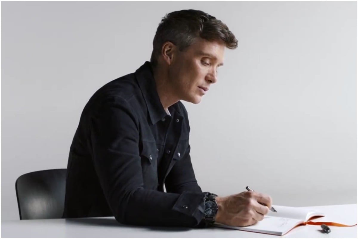 Cillian Murphy sits at a desk and writes in a notebook.