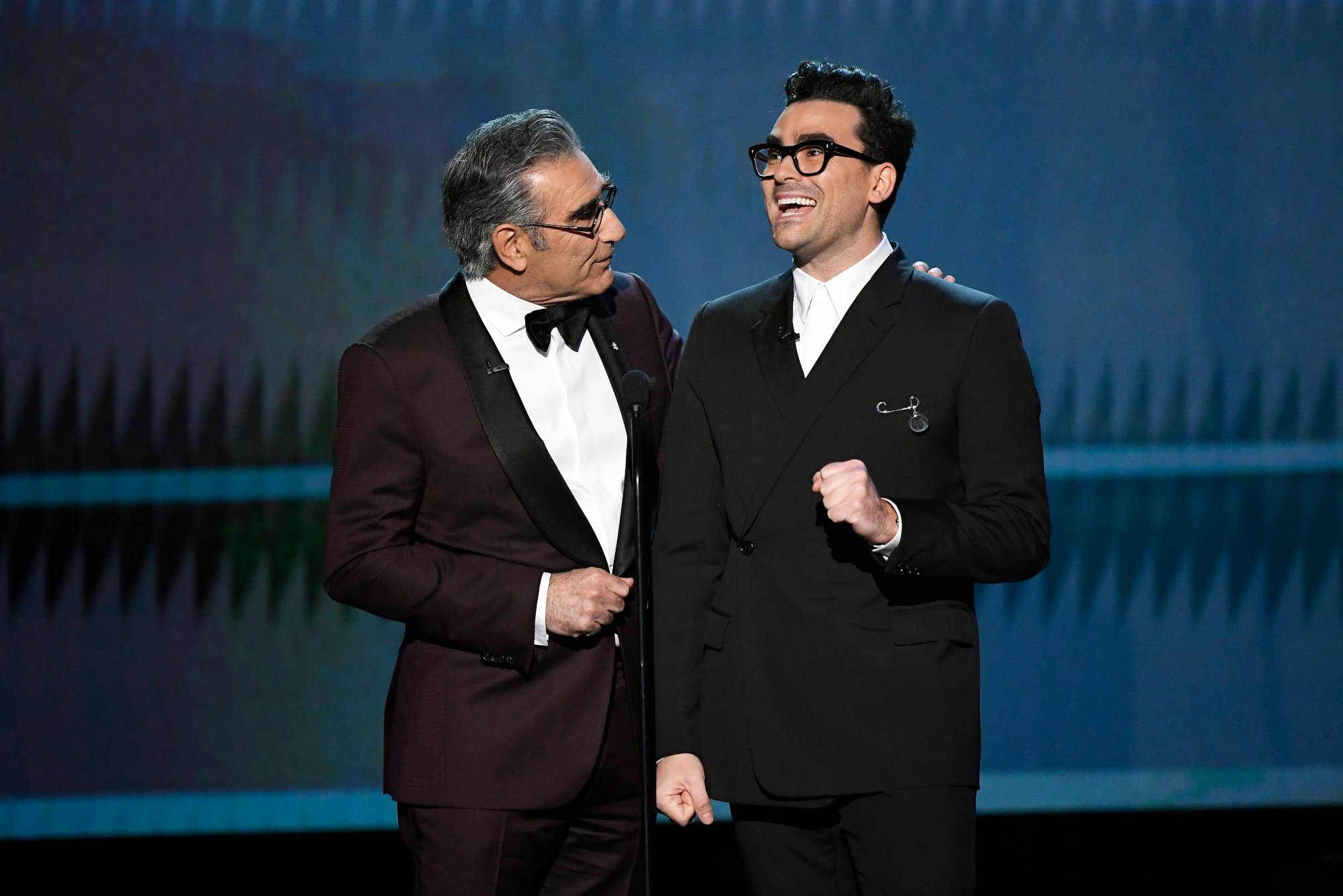 Schitts Creek stars Dan Levy and Eugene Levy at an awards show. Eugene, right, has his arm around his son and is looking at him. Dan is holding an envelope and laughing.
