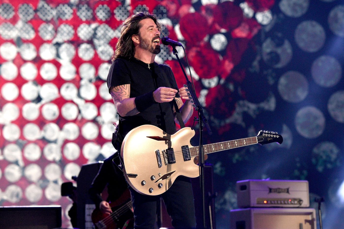 Dave Grohl from the Foo Fighters on stage playing a guitar in a pair of black pants and a black T-shirt in front of a background with red, white, and blue circles.