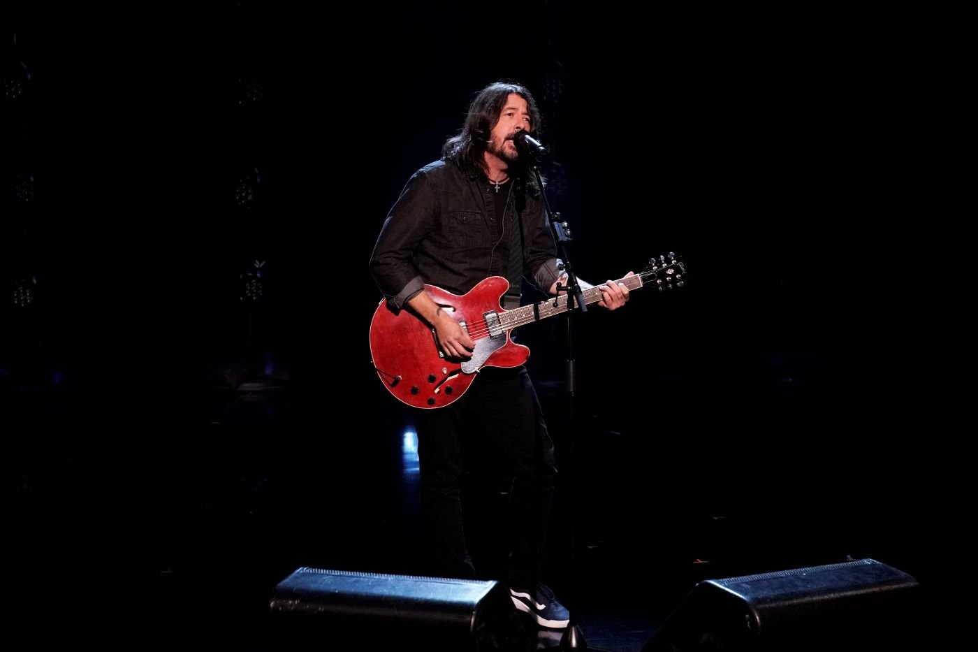 Dave Grohl with his guitar dressed all in black standing in front of a black background and stage with a spotlight on him.