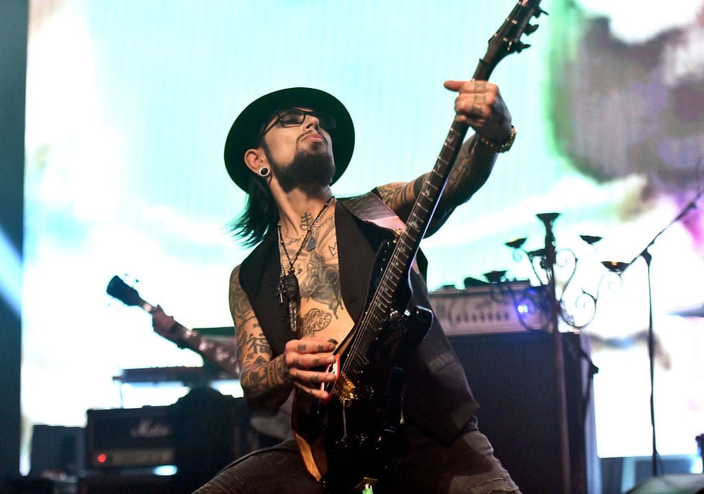 Dave Navarro plays guitar on stage in a black open vest, a black hat, and black jeans.