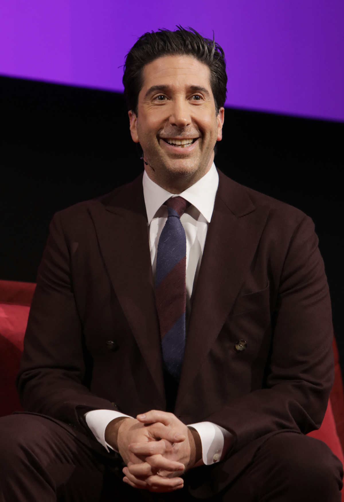 David Schwimmer wearing a suit and tie, seating with his hands clasped and smiling.