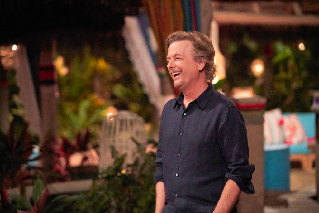 David Spade, wearing a black button-up shirt, stands laughing during hosting duties.