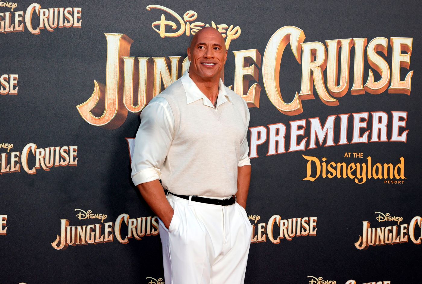 Dawayne Johnson standing in front of a black backdrop with Jungle Cruise written in gold in various places dressed in a pair of white pants with a cream sweater with a white collar and black belt.