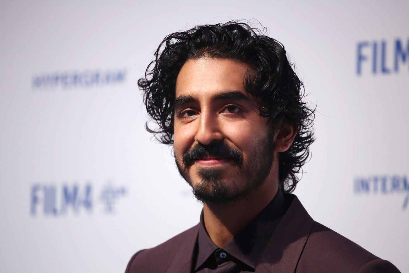 Dev Patel wearing a brown suit with a brown shirt in front of a white and blue background.