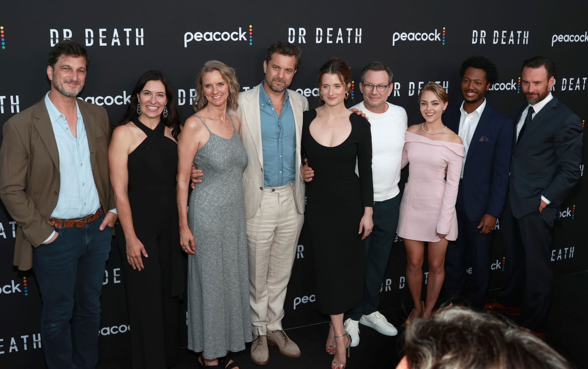 Nine cast members from 'Dr. Death' are dressed professionally standing in front of a black background with Dr. Death and Peacock written on it.