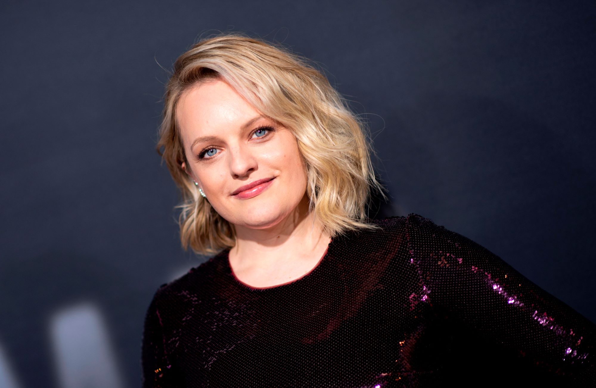 Elisabeth Moss standing in front of black background with a dark purple top.