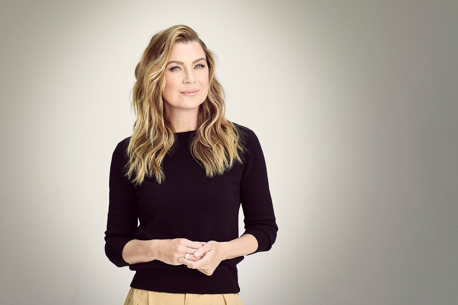 Grey's Anatomy star Ellen Pompeo wearing a black top and beige pants and wearing her blonde hair down. She's standing in front of a solid grey background and staring at the camera.