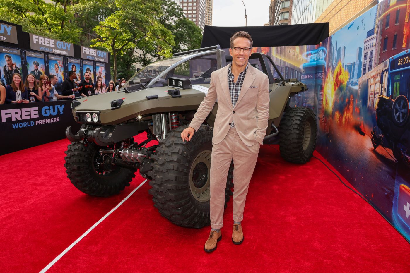 Ryan Reynolds standing in front of an army green truck on the red carpet at a movie premiere wearing a tan suit with a blue undershirt. 