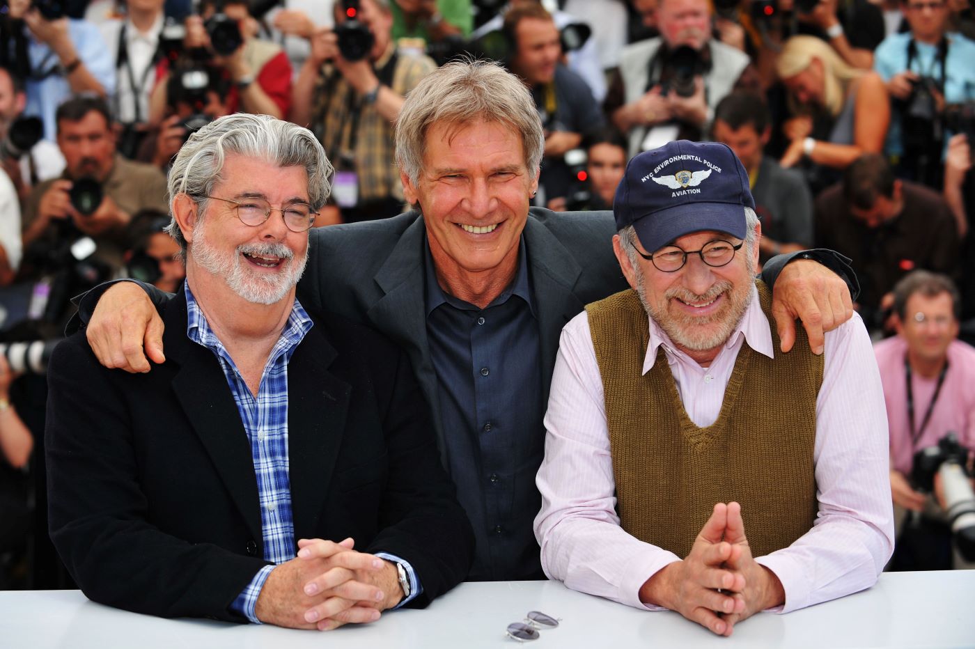 George Lucas, Harrison Ford, and Steven Spielberg sitting behind a white desk dressed professional in front of a blurred audience. Collaborators of 'Indiana Jones' and 'Star Wars'