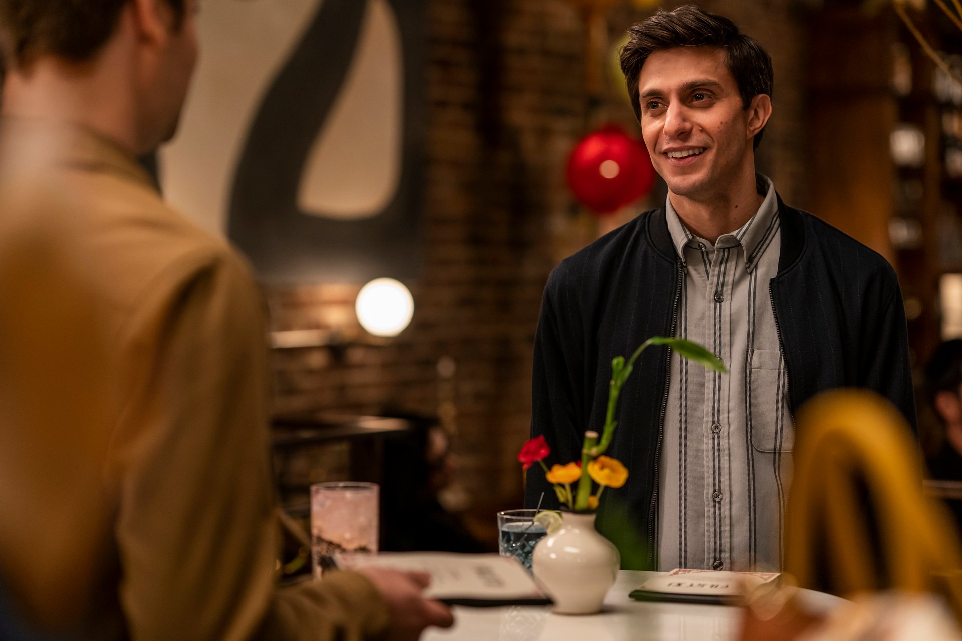 Gideon Glick wears a striped shirt and jacket in a bar in 'The Other Two.'