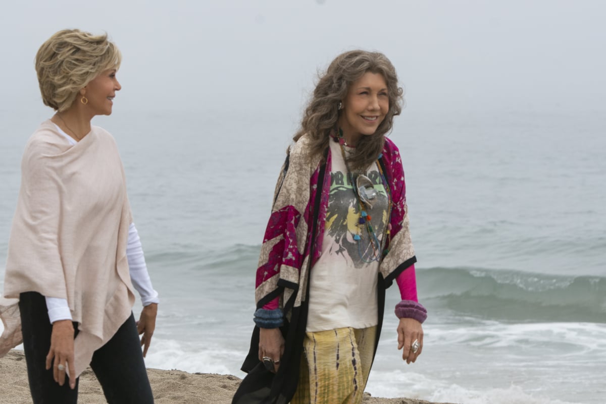 Grace and Frankie smile and walk along the beach.