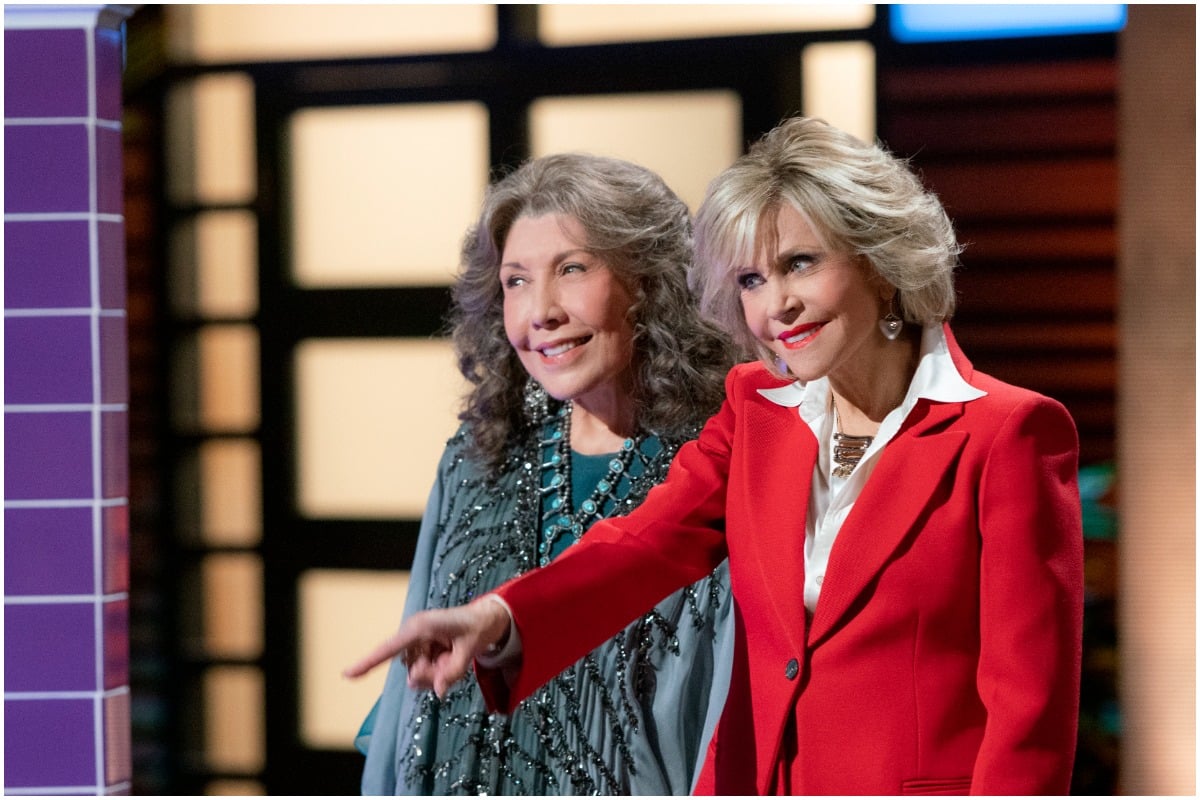 Grace and Frankie pitch a business idea. Grace wears a red suit and Frankie wears a blue parka.