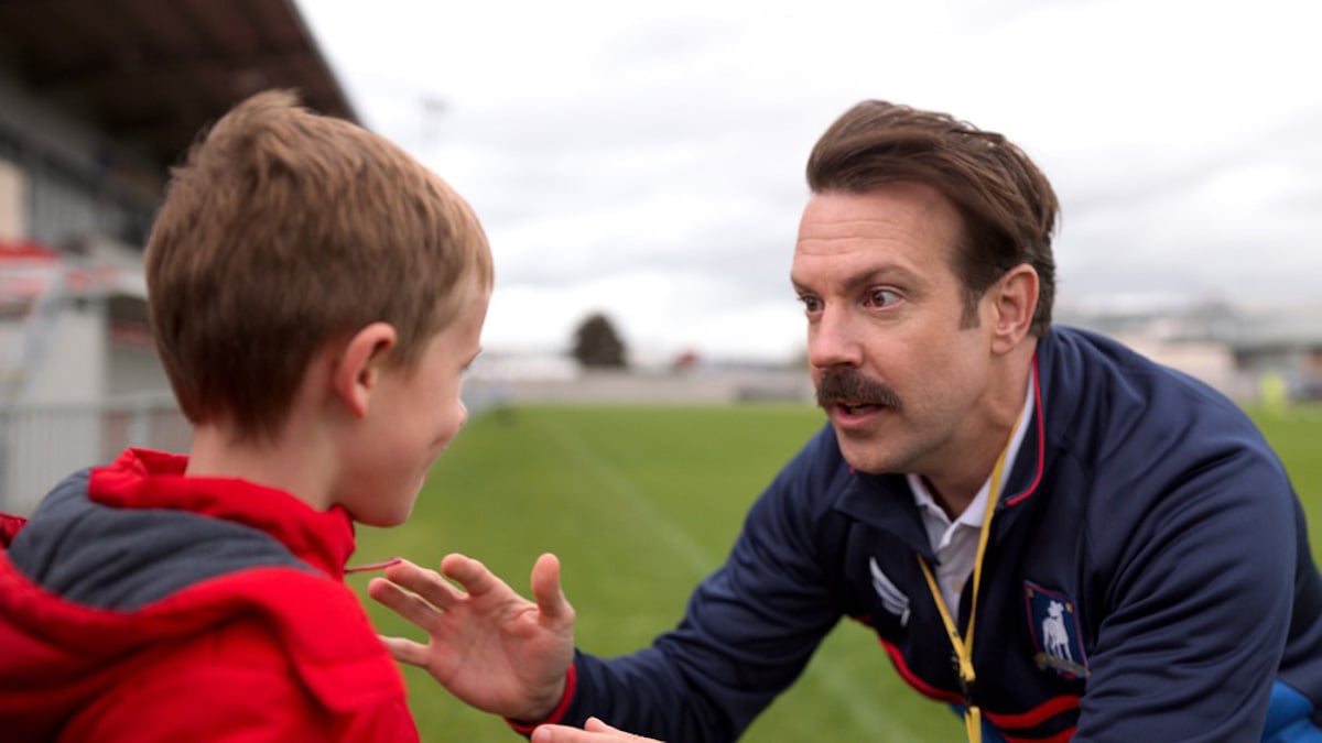 Jason Sudeikis wears a blue shirt and talks to Gus Turner on a soccer field in 'Ted Lasso' Season 1