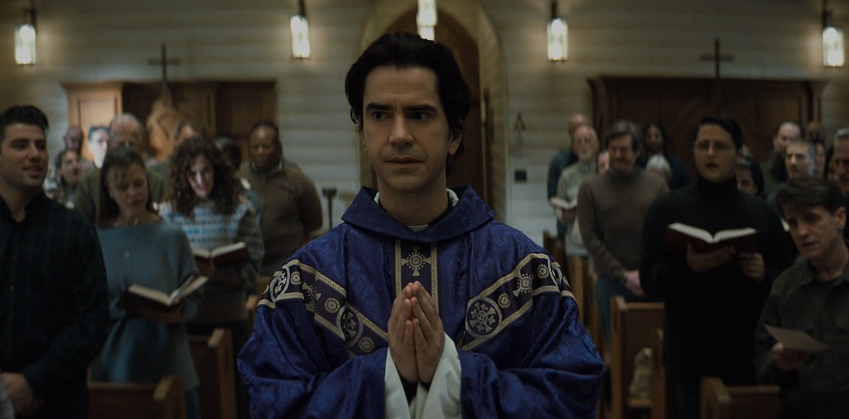 Hamish Linklater wears blue priest's robes and walks down a church aisle in 'Midnight Mass.'