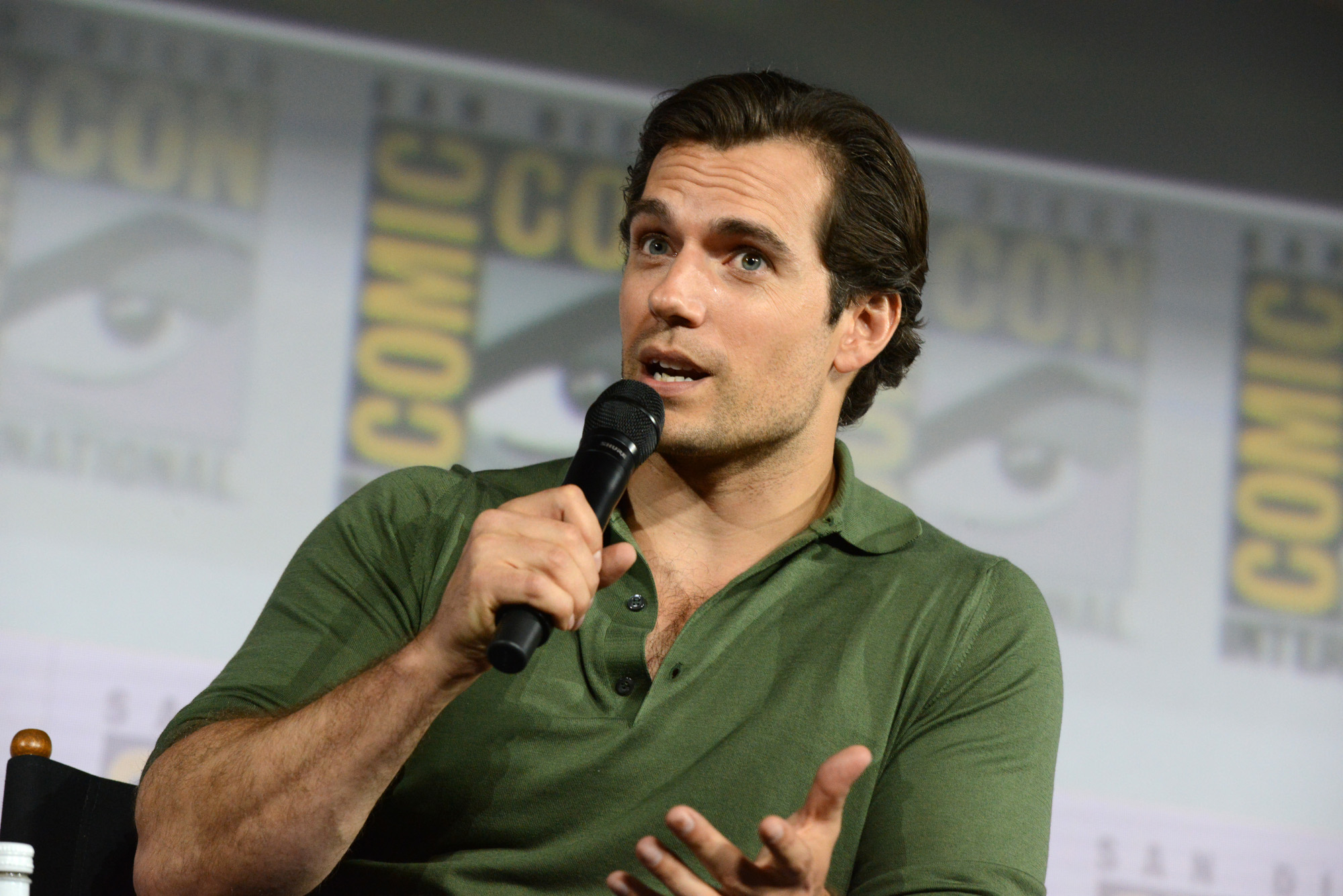 Henry Cavill, who almost played James Bond, wearing a great polo shirt and holding a microphone on a Comic-Con panel. He's sitting and there is a Comic-Con wall behind him.
