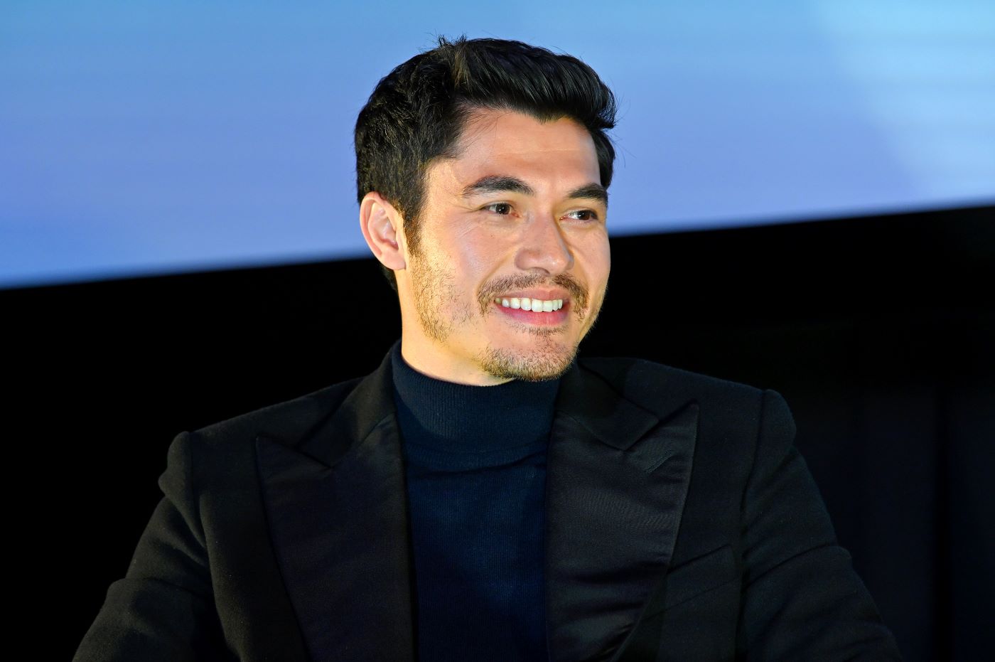 Henry Golding is wearing a black suit jacket and a black undershirt against a black and light blue background.
