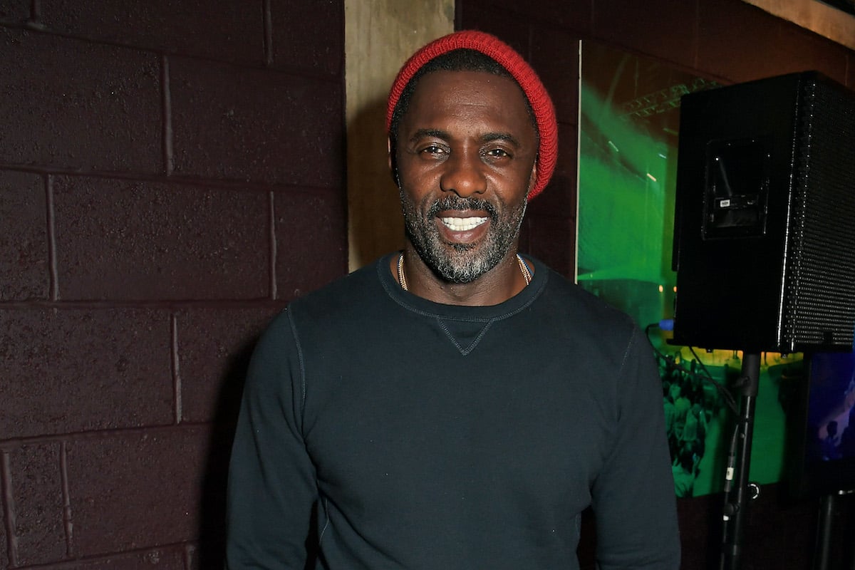 Idris Elba wears a black shirt and red hat as he smiles and poses