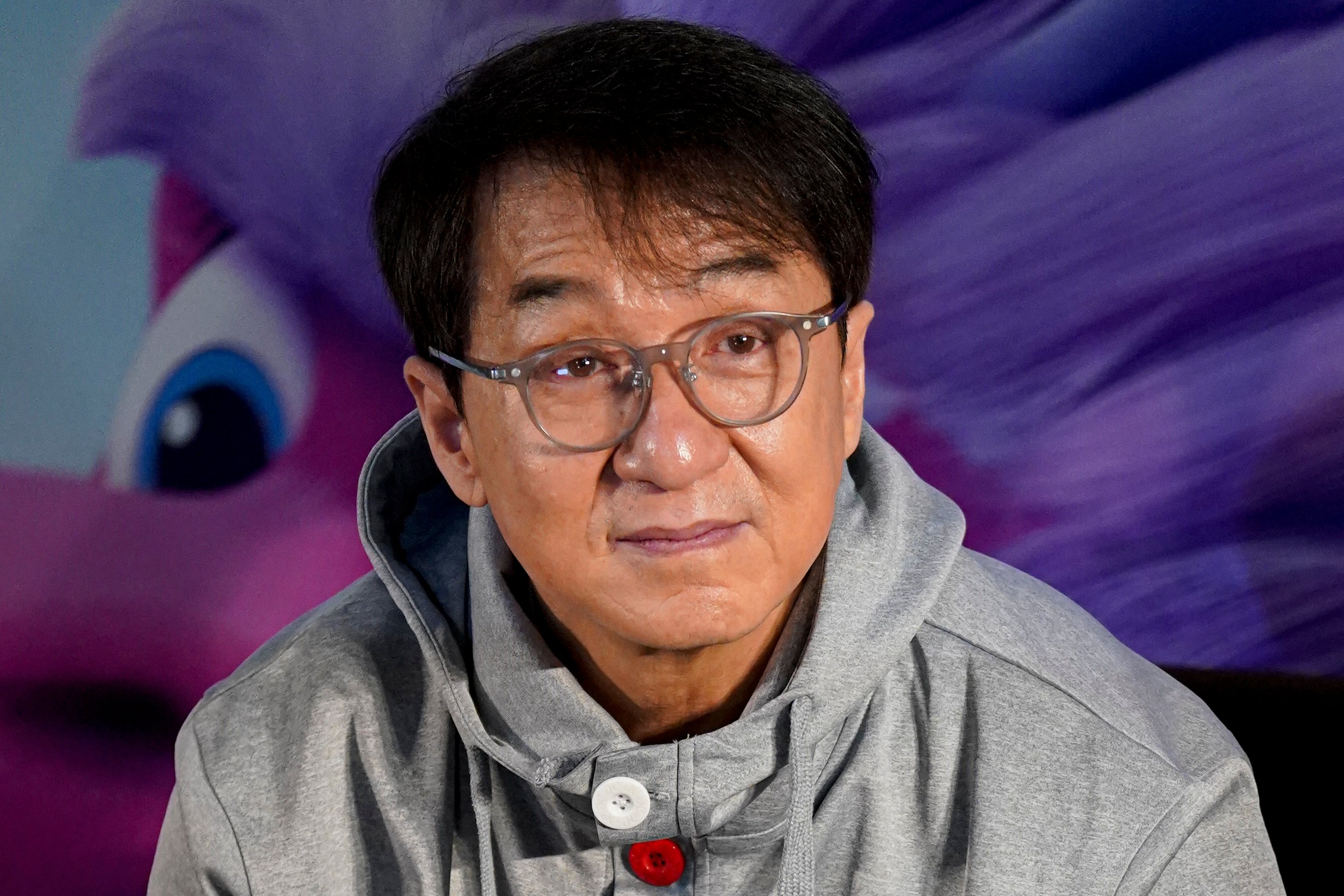 Jackie Chan at press event wearing gray hooded sweater