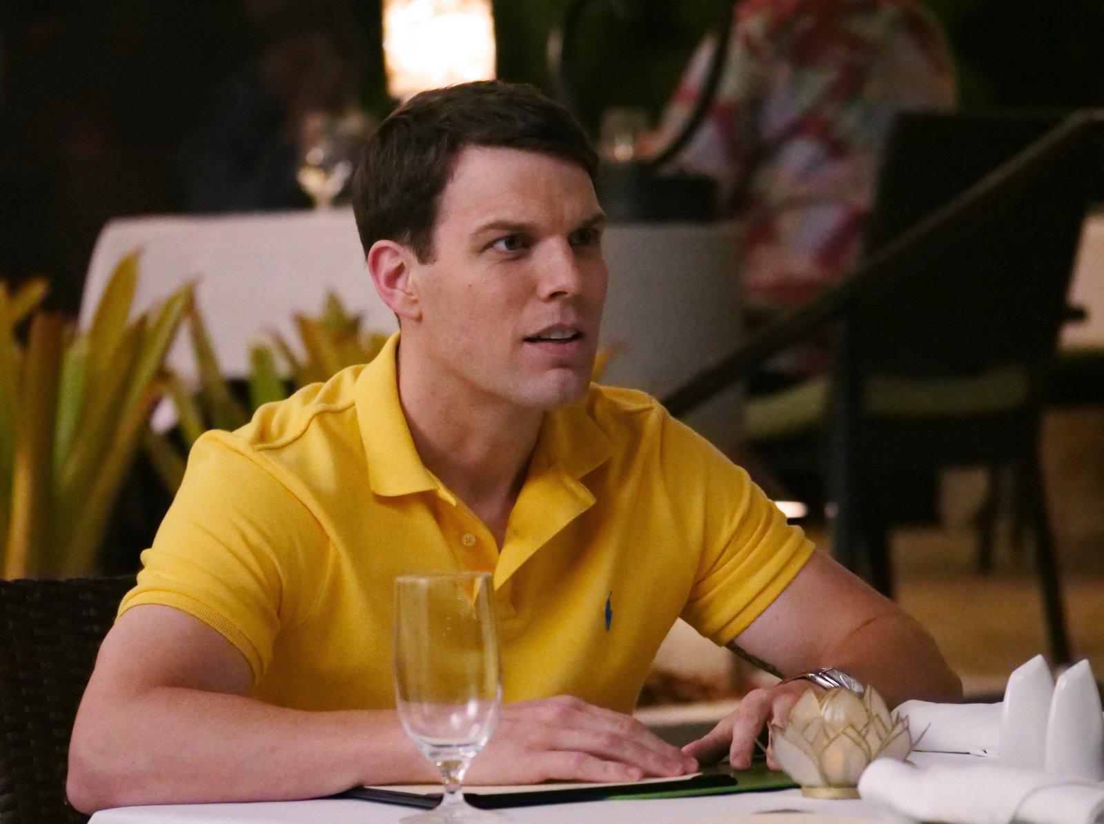 Jake Lacy as Shane in HBO's 'The White Lotus' finale. He's wearing a yellow polo shirt and leaning on a dinner table, which has a white table cloth and is set with utensils and glasses.