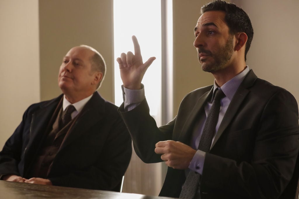 James Spader as Raymond 'Red' Reddington sits next to Amir Arison as Aram Mojtabai who is signing with ASL. They are both wearing dark suits and ties.