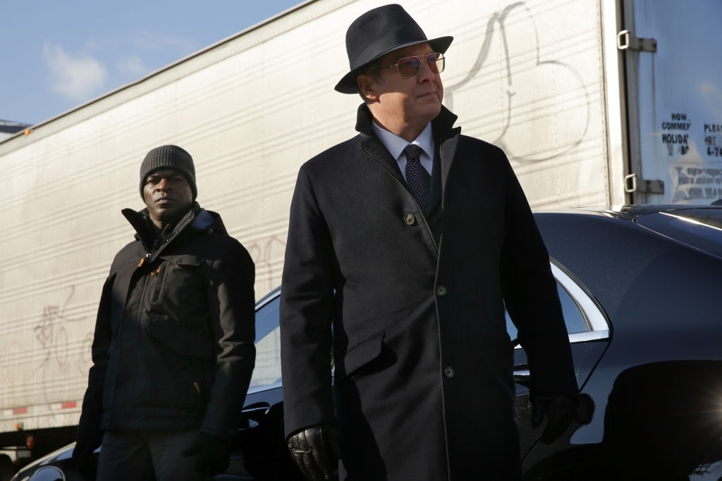 Hisham Tawfiq as Dembe Zuma stands outside next to James Spader as Raymond 'Red' Reddington. Both men are dressed in suits and overcoats. 