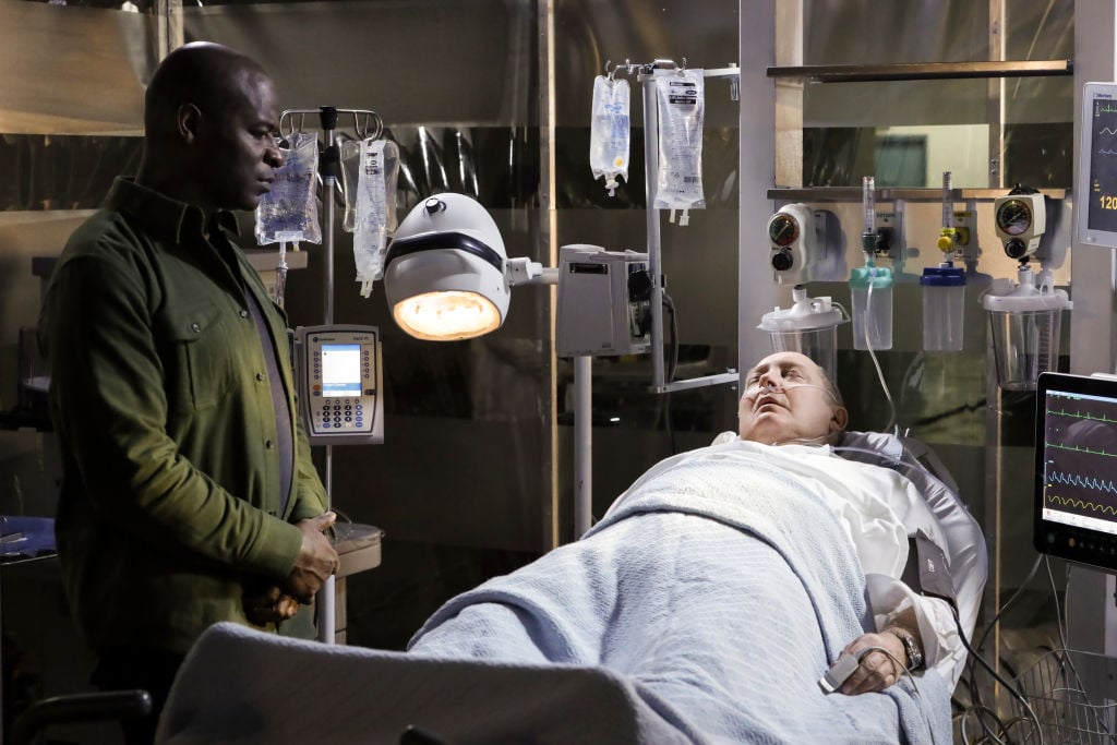 Hisham Tawfiq as Dembe Zuma stands next to an ailing James Spader as Raymond 'Red' Reddington who lies in a hospital bed connected to machines.