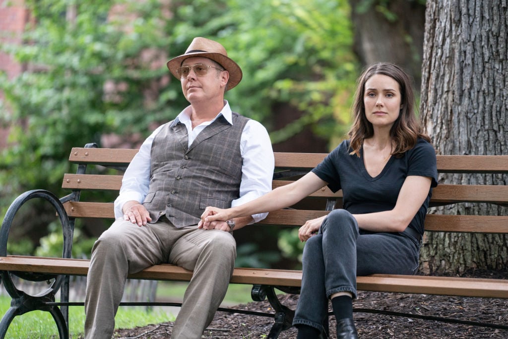 James Spader as Raymond 'Red' Reddington, Megan Boone as Elizabeth Keen sit on a bench outside together, holding hands.