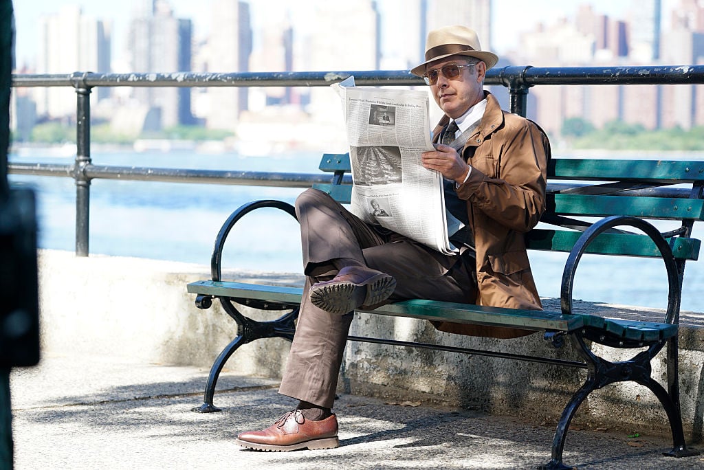 James Spader as Raymond 'Red' Reddington  sits on a bench while reading a newspaper. He's dressed in a brown suit, fedora, and sunglasses.
