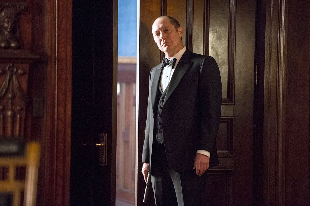 James Spader as Raymond 'Red' Reddington  stands in a dark suit with a gun in his right hand, at his side.