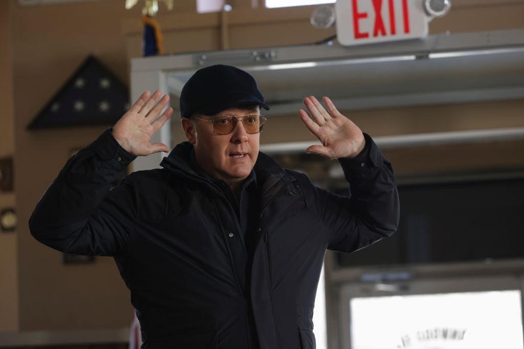 James Spader as Raymond 'Red' Reddington stands with his hands up to surrender. He's wearing a dark coat and hat and tinted glasses.