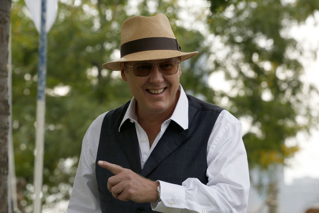 James Spader as Raymond 'Red' Reddington  smiles for the camera. He's wearing a tan fedora, dark gray vest, sunglasses, and a long-sleeve white shirt.