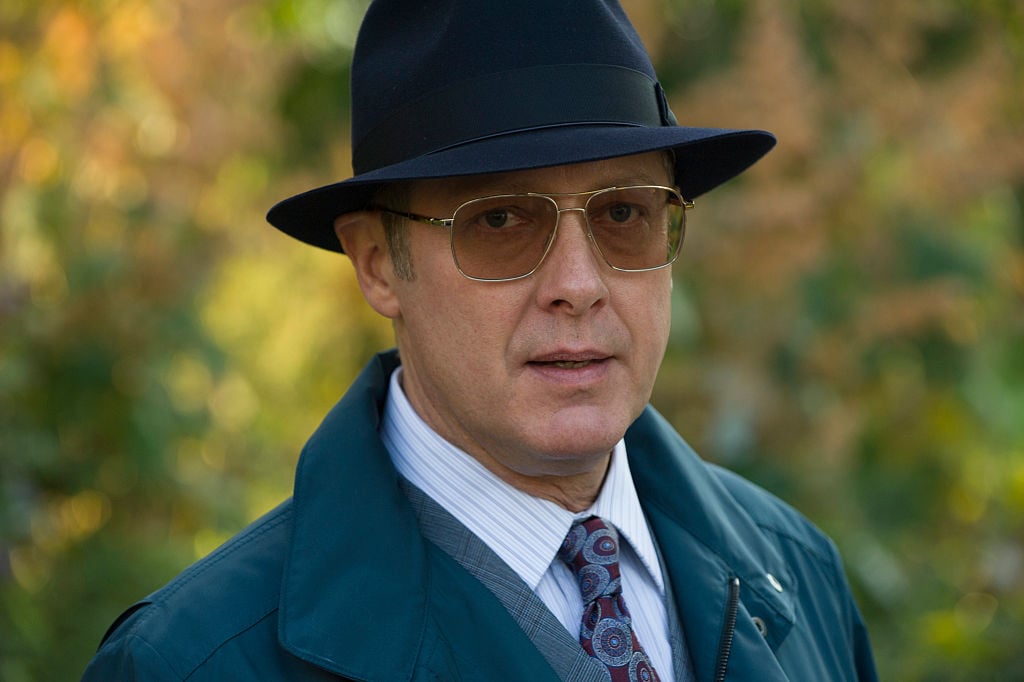 James Spader as Raymond 'Red' Reddington is dressed in a navy trench coat, matching fedora, and sunglasses as he looks directly into the camera for a close-up shot.