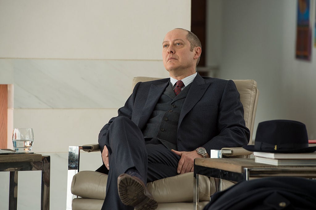 James Spader as Raymond 'Red' Reddington is dressed in a dark suit as he sits in a chair, unimpressed.