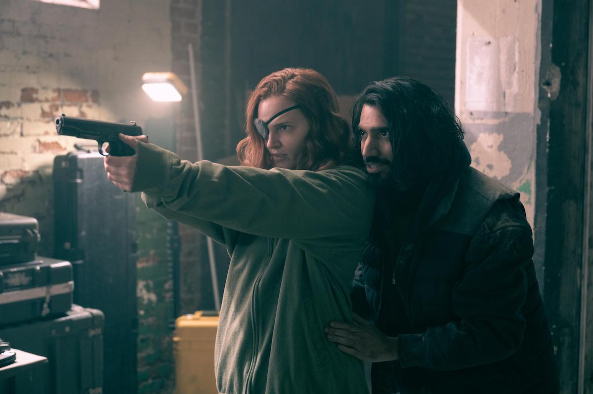 Madeline Brewer and Omar Maskati as Janine and Steven in 'The Handmaid's Tale' Season 4. In the photo, Brewer wears a grey zip-up hoodie and a beanie and an eyepatch as she points a gun. Maskati stands behind her, his hands on her waist. He wears a jacket with a vest over it. They're in a dilapidated warehouse where the Chicago resistance against Gilead hides. In 'The Handmaid's Tale' Season 4, Janine's backstory was revealed. Brewer says Janine's backstory is in the back of her mind in every scene she does in the Hulu series.