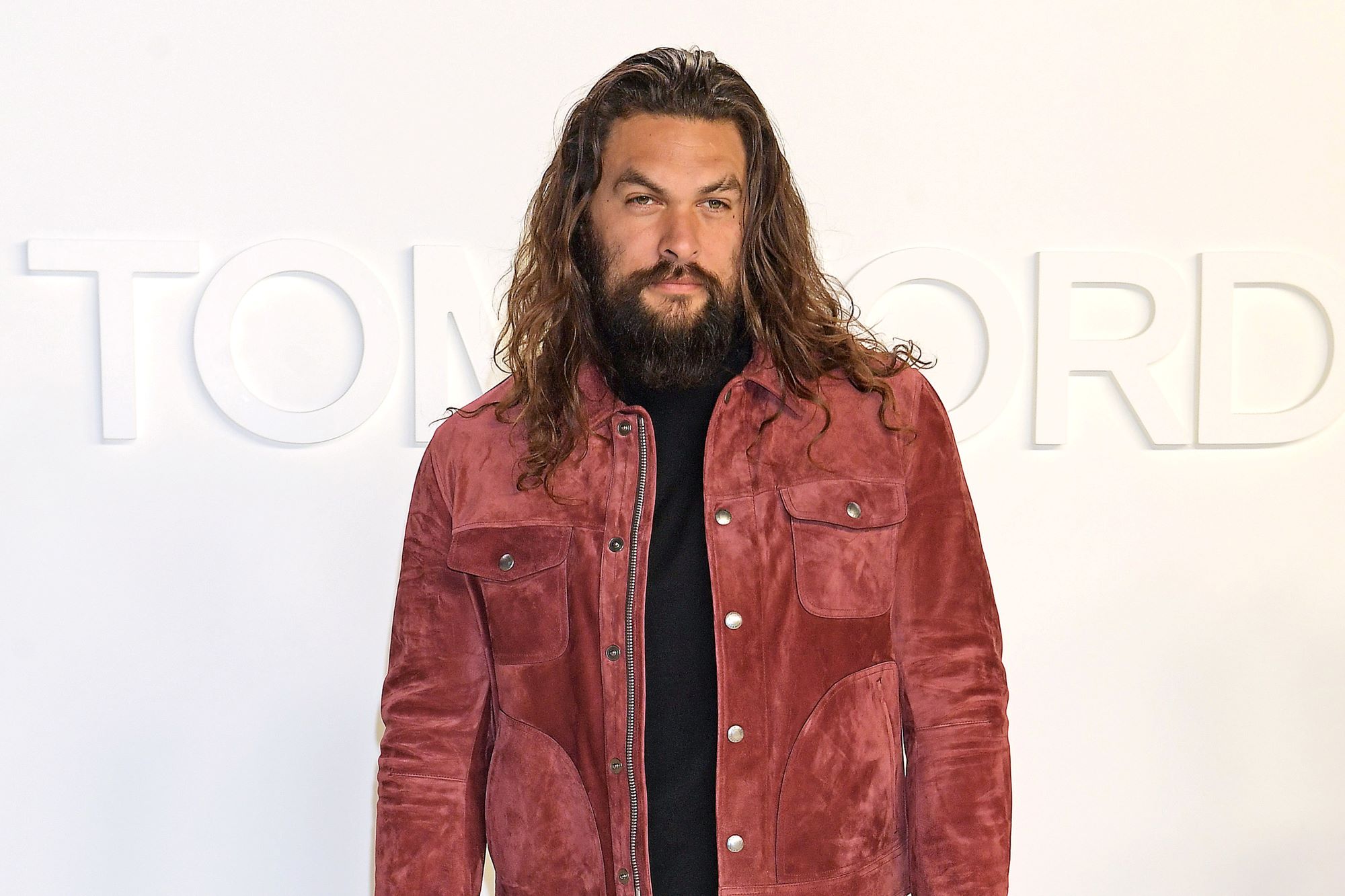 Jason Momoa dressed in a redish brown jacket with a black shirt and pants in front of a white background.