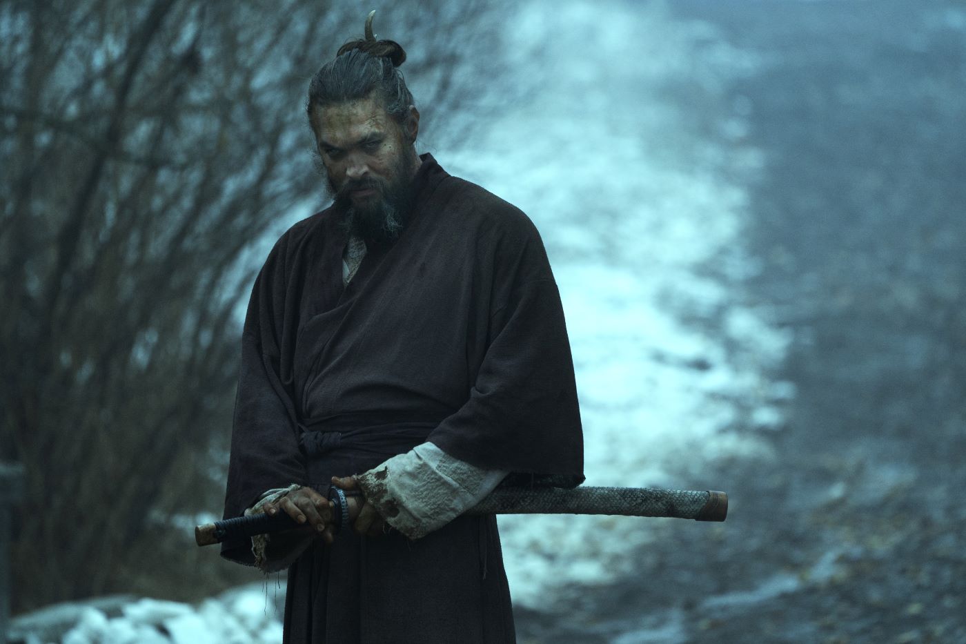 Jason Momoa in a scene from 'See' standing in a wooded snowy area with a brown robe and a blood stained white shirt sleeve underneath with his hand on his sword about to draw it.