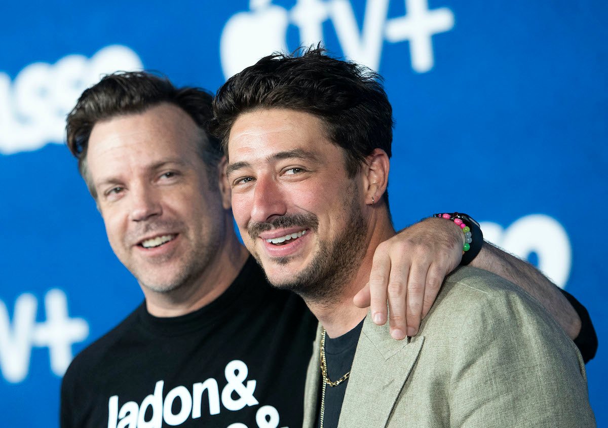 Jason Sudeikis (L) and Marcus Mumford (R) at the 'Ted Lasso' Season 2 premiere on July 15, 2021. Sudeikis wears a black long-sleeved shirt with white writing and Mumford wears a beige suit jacket. They stand in front of a blue backdrop. Sudeikis has his left arm around Mumford's shoulder.