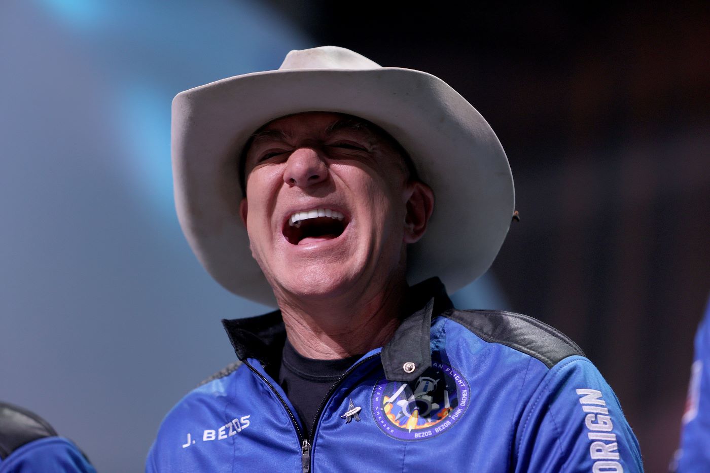 Jeff Bezzos in his Blue Origin space suit and cowboy hat standing in front of a blue and black background.