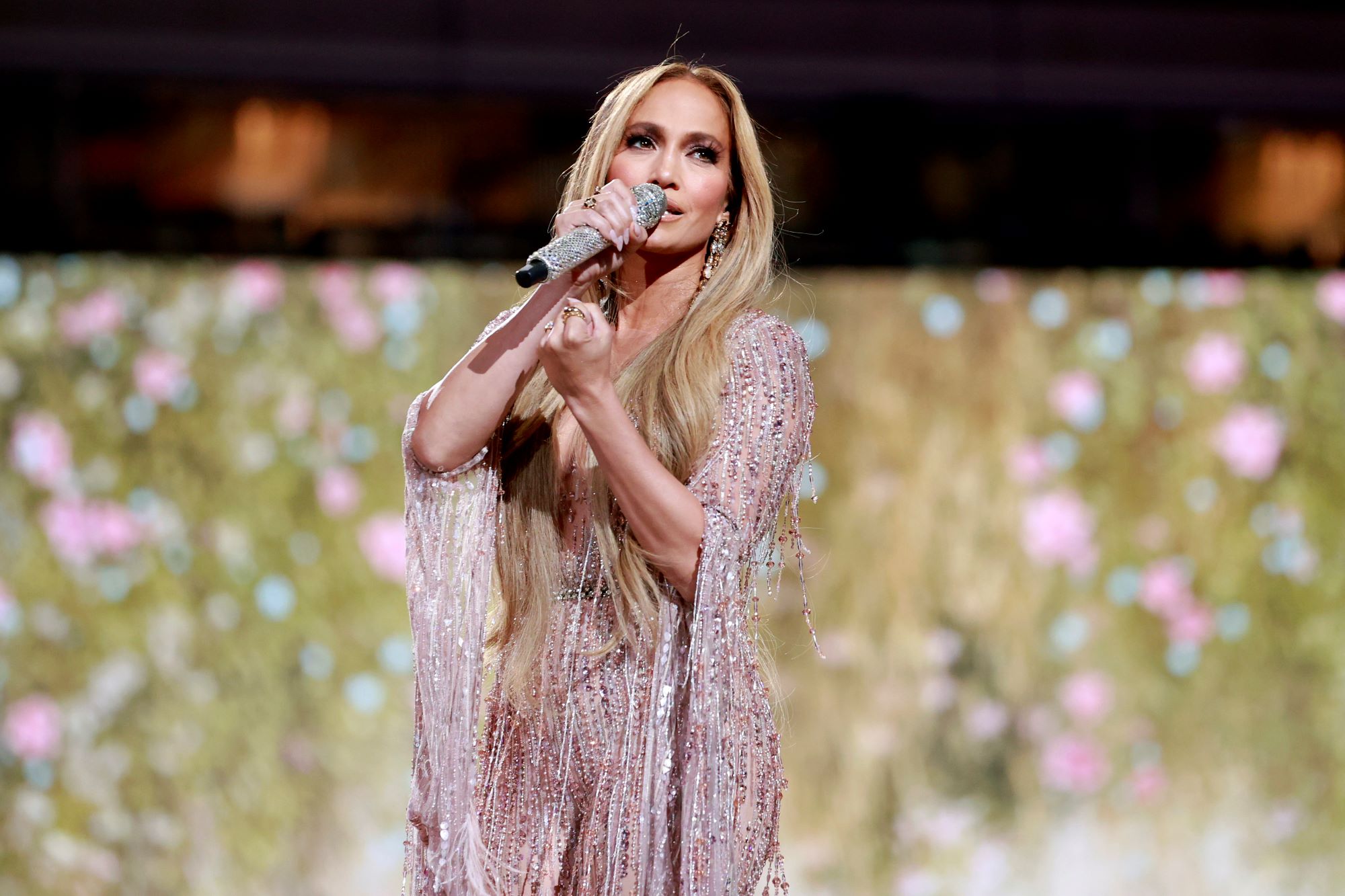 Jennifer Lopez dressed in a champagne colored sparkly dress with a similar sparkly background with green mixed in.