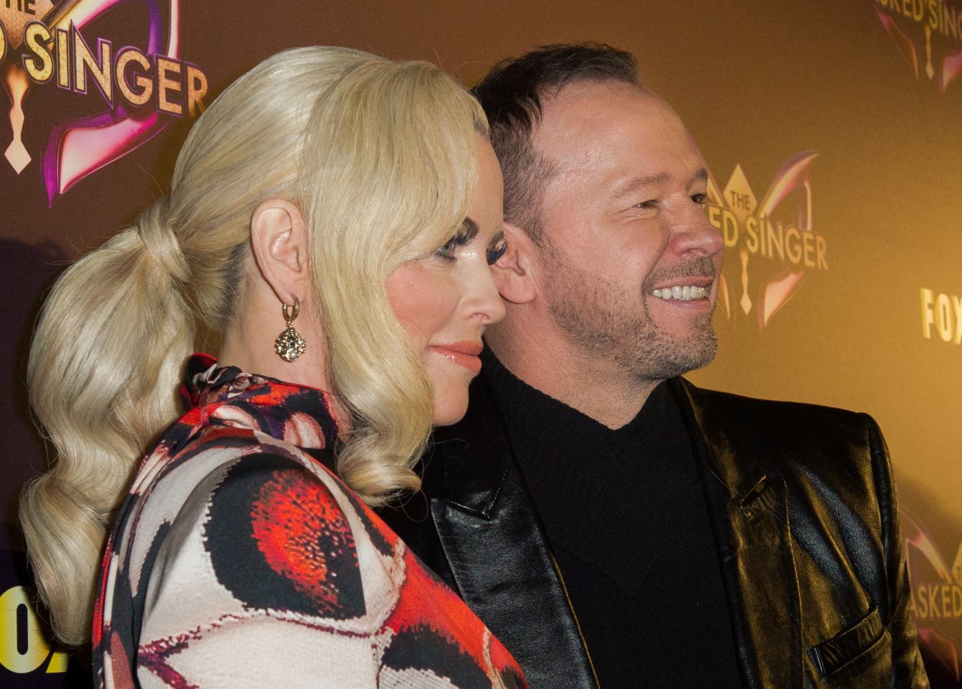 Jenny McCarthy-Wahlberg and Donnie Wahlberg standing in front of a black background with 'The Masked Singer' logo on it dressed in casual-professional clothing.