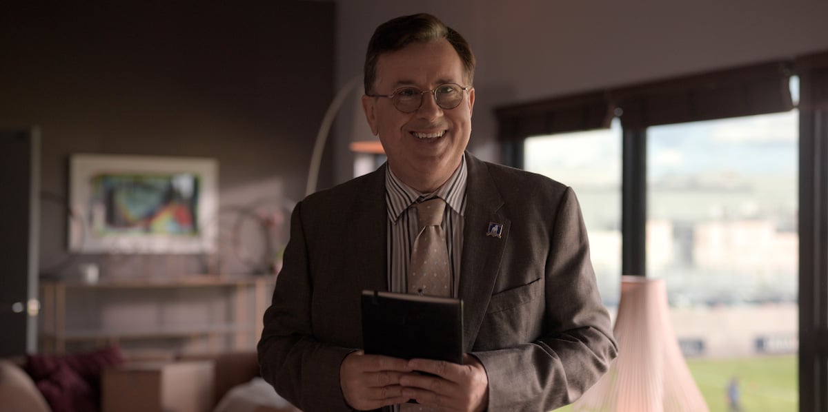 Jeremy Swift smiles wearing a suit and tie while holding a notebook as Leslie Higgins in 'Ted Lasso'