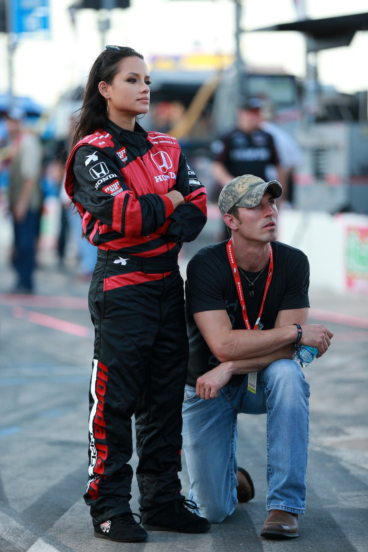 Jessica Graf stands in racing attire, looking off into the distance, while husband Cody Nickson kneels by her side.