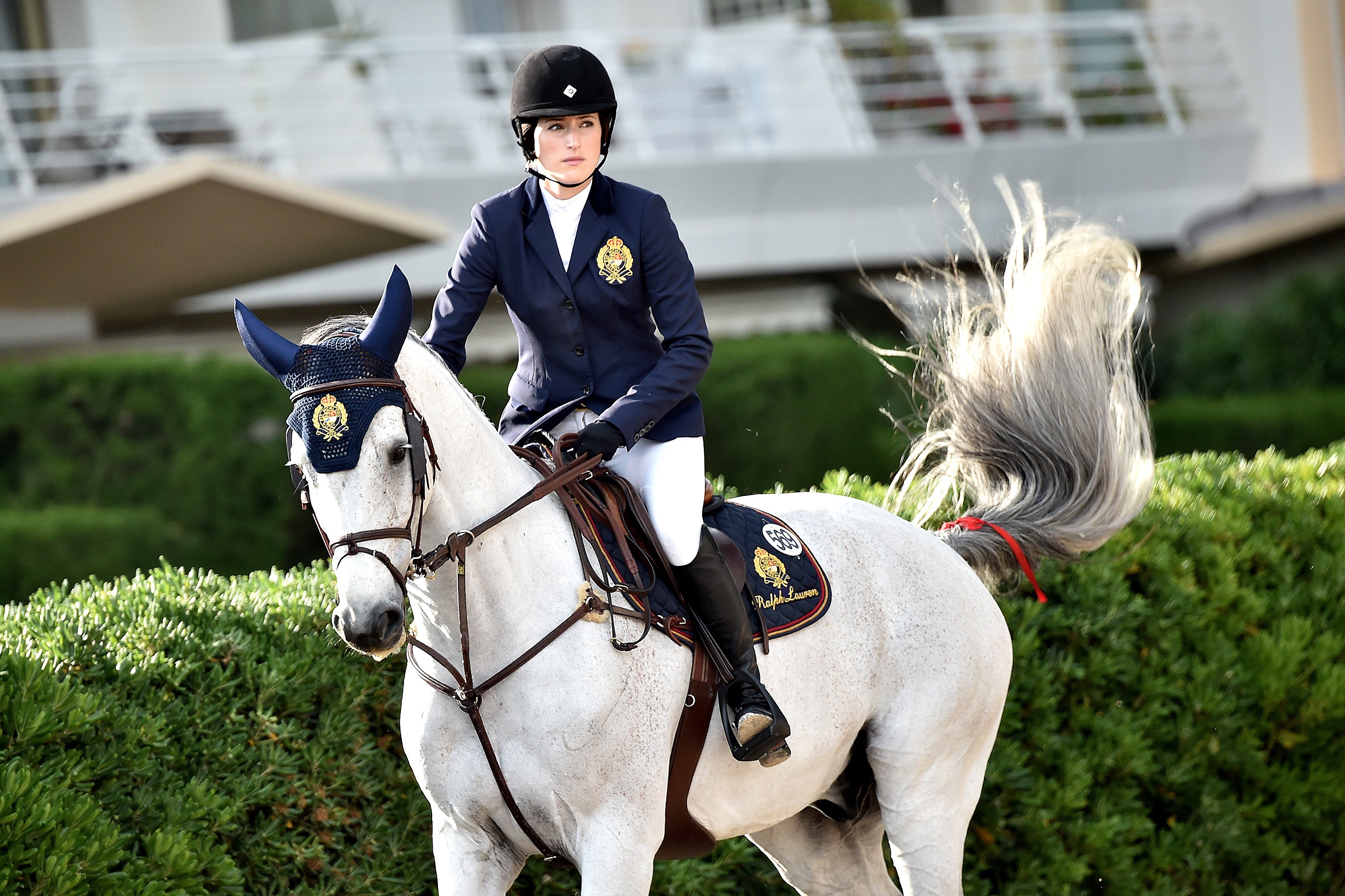 Jessica Springsteen rides a white horse while wearing a helmet and blue jacket.