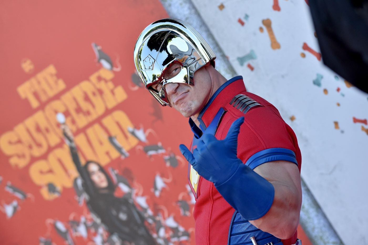 John Cena dressed as a DC superhero, Peacemaker, standing in front of a background of a Suicide Squad poster with confetti.