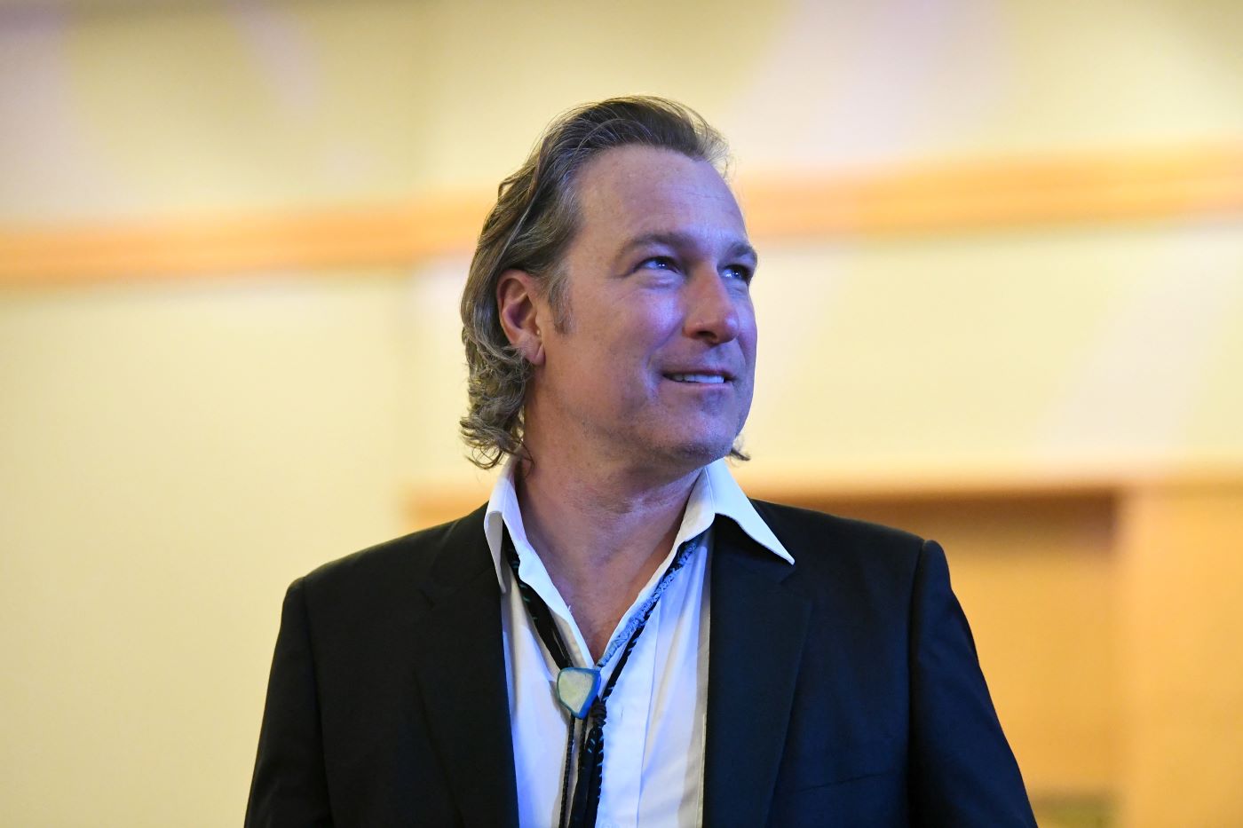 John Corbett is dressed in a black suit jacket and a white undershirt standing in front of a cream background