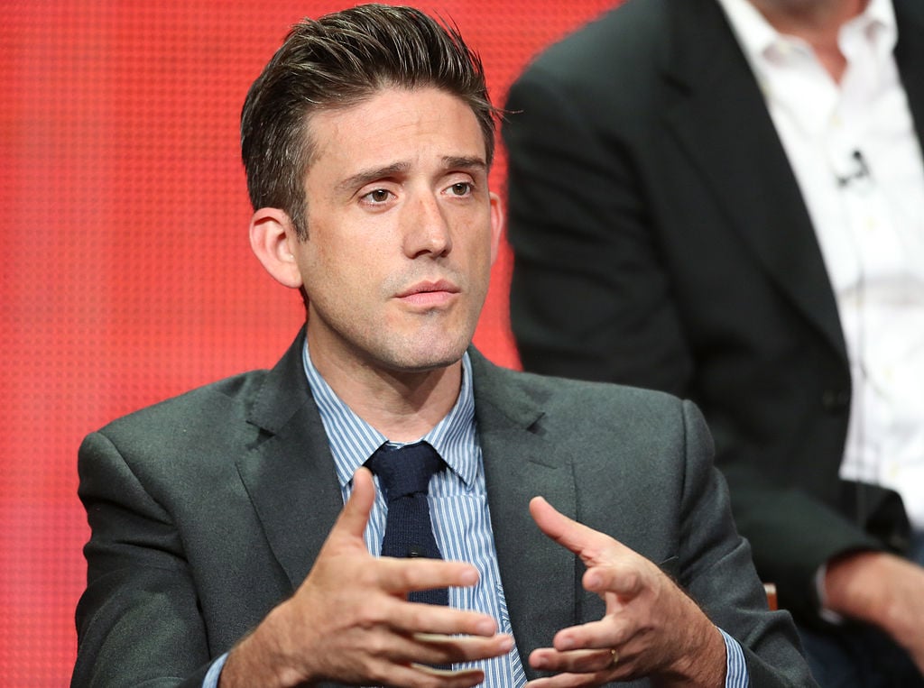 Jon Bokenkamp speaks during a panel. He's dressed in a grey suit and sits next to 'The Blacklist' cast members.