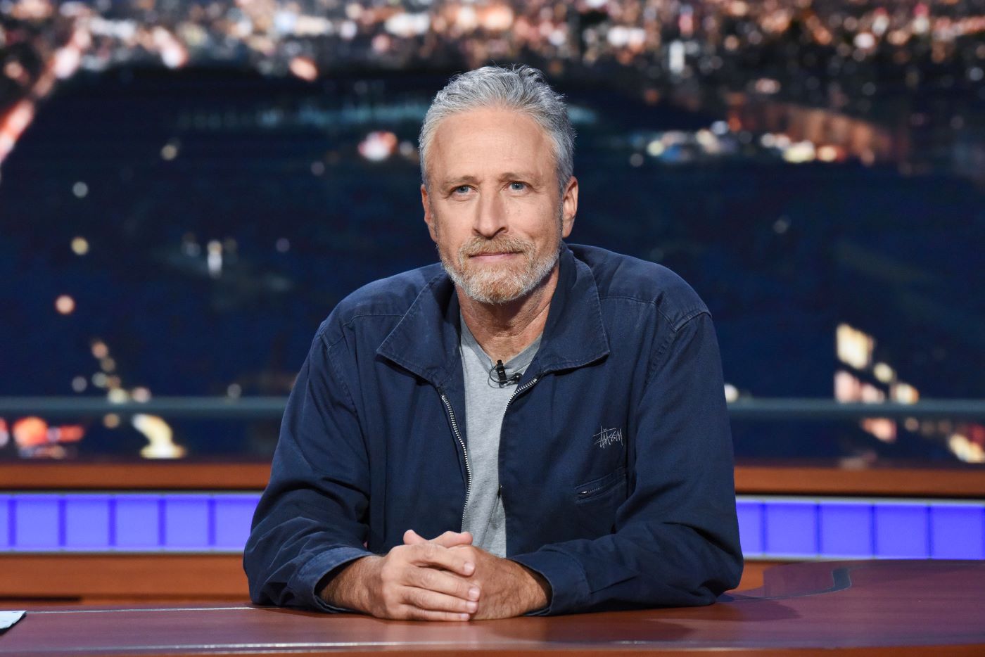 Jon Stewart sitting behind a desk with a city in the background wearing a grey shirt with a blue jacket.